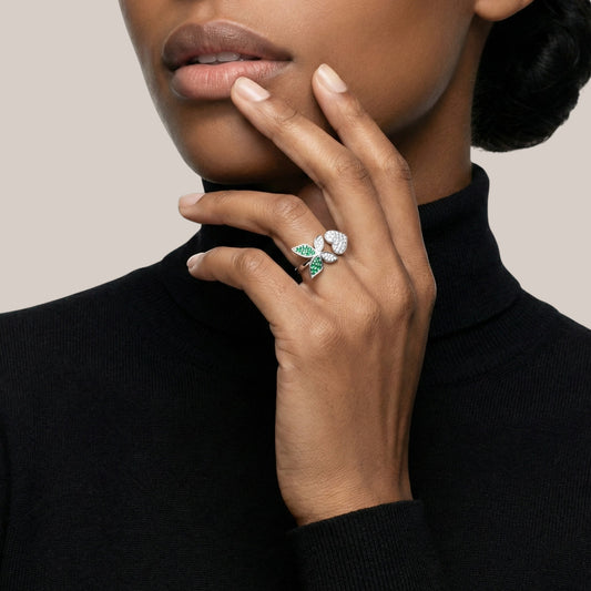 Close-up of a hand wearing a ring with green gemstone and diamonds, against a neutral background.