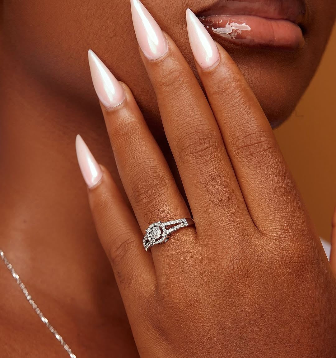 A person showcasing their engagement ring with a round cut diamond on their finger. The ring has a gold band and the person's nails are painted in a light color.