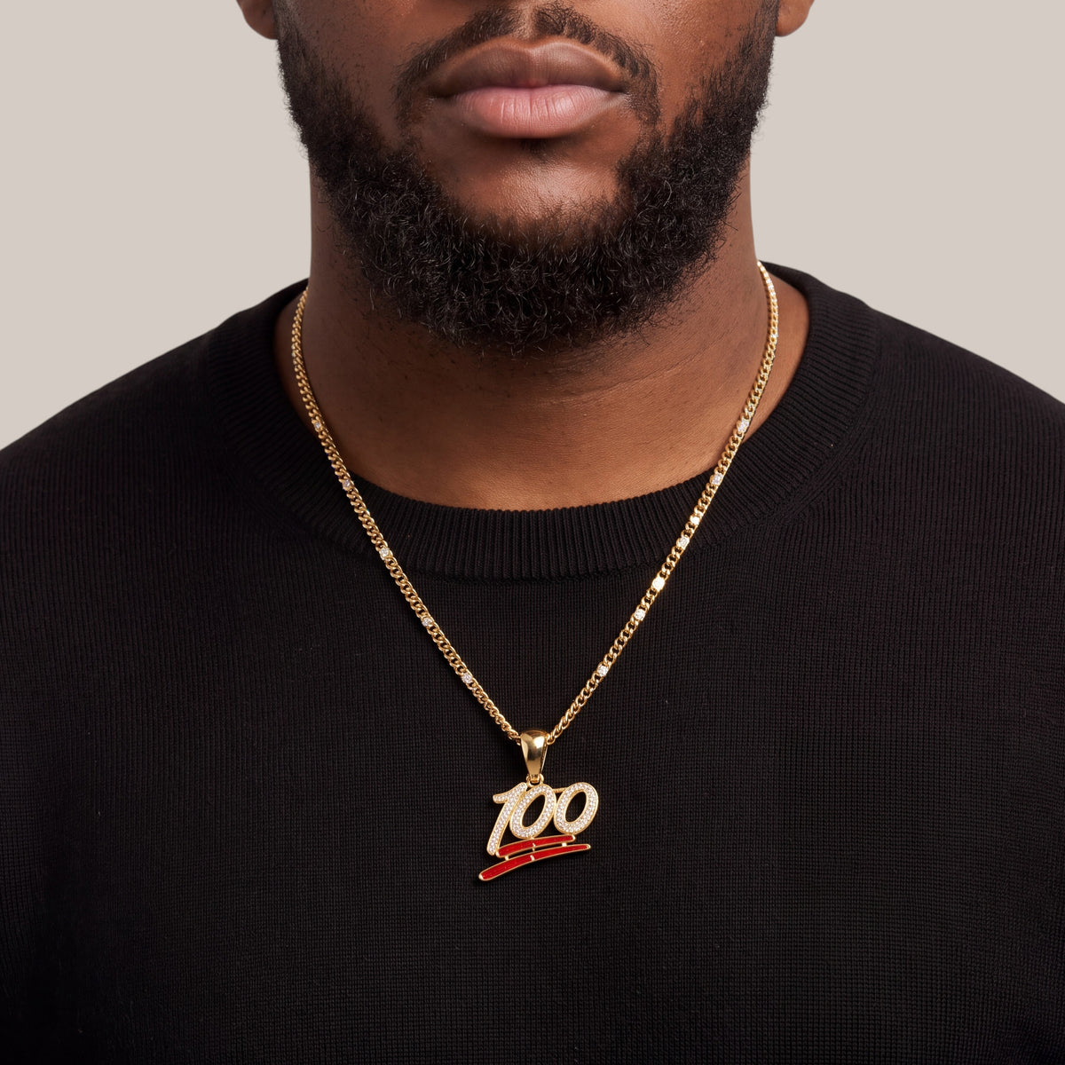 A close-up of a man wearing a gold chain necklace with a '100' pendant featuring a red accent line, styled with a black crewneck sweater against a neutral beige background.