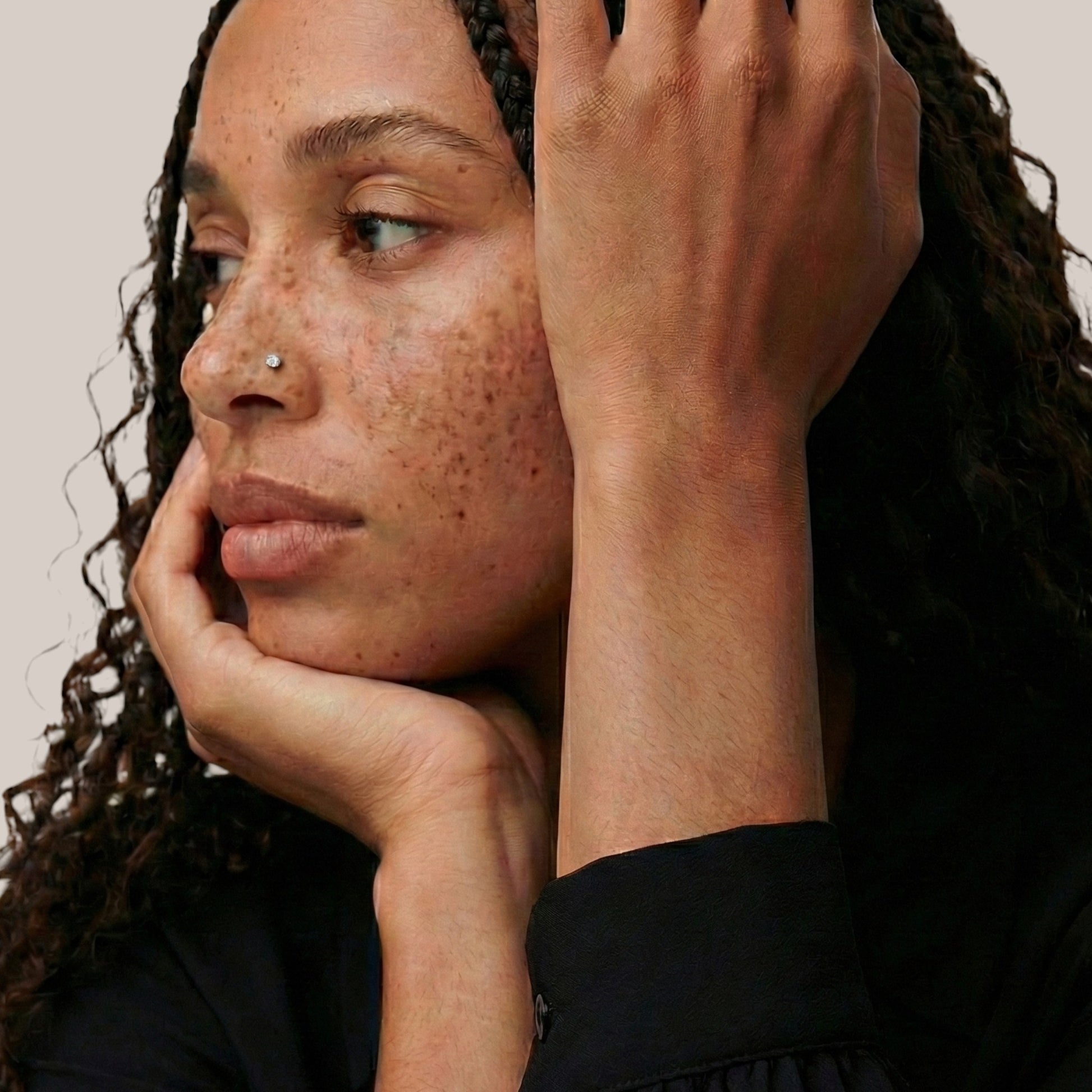 Woman with freckles and a nose piercing, touching her face against a neutral background