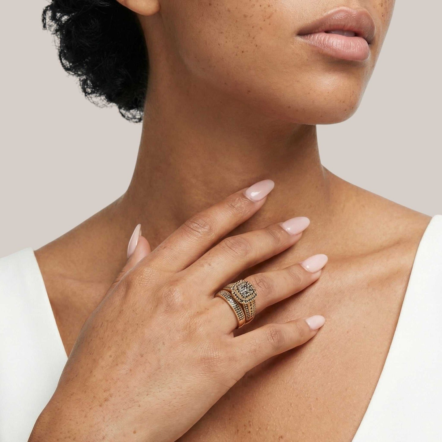 Close-up of a woman's hand wearing a gold ring with a diamond, touching her neck against a neutral background.