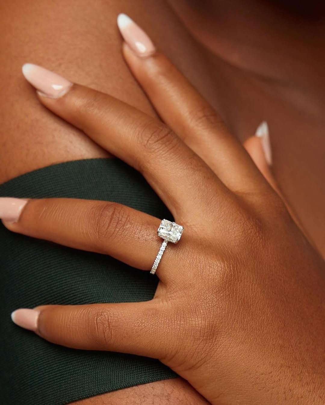 A person's hand displaying a radiant cut moissanite engagement ring on a dark green velvet surface.