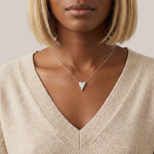 Woman wearing a beige sweater and silver heart necklace against a neutral background