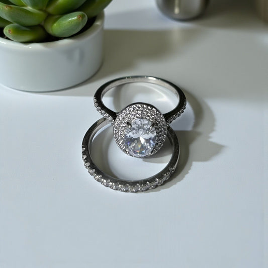 Silver ring with a large central gemstone on a light surface, with a plant in the background.