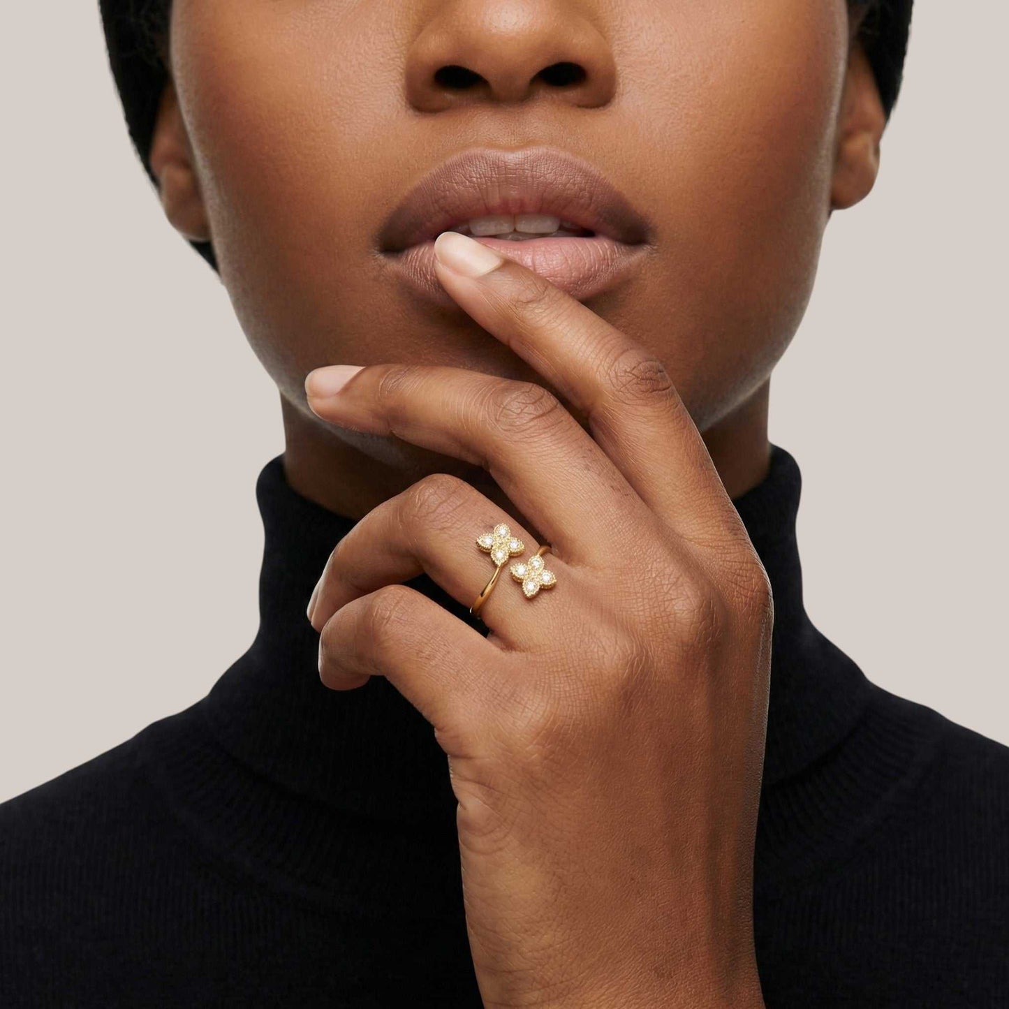 Close-up of a person wearing a gold ring with three small stones, touching their lips against a neutral background.