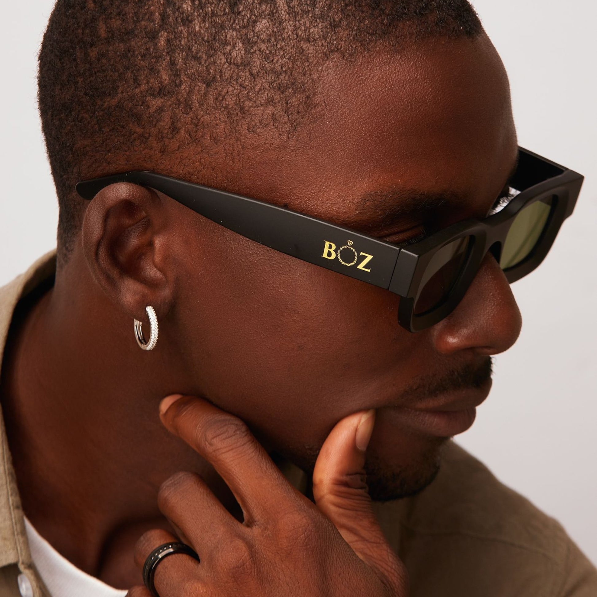 Person wearing black sunglasses with 'BOZ' logo, touching their chin against a neutral background